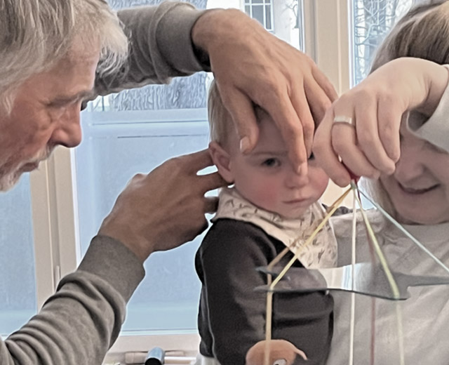 Ein Osteopath behandelt ein Kleinkind am Kopf, das Kind lagert auf dem Arm der Mutter und schaut ein Mobile an